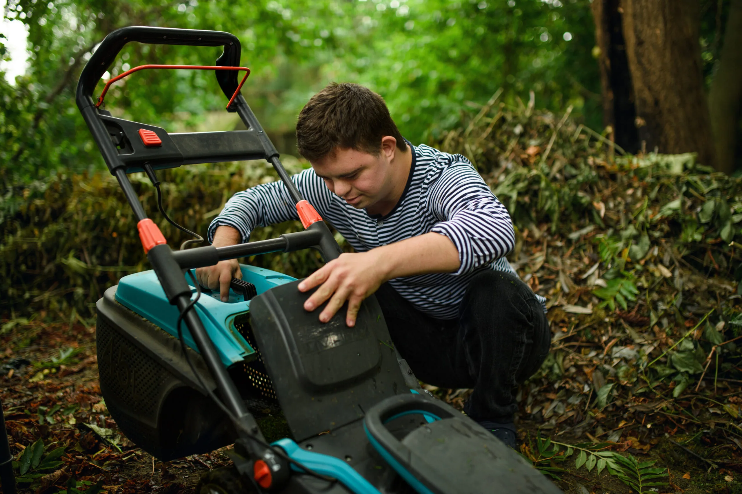 Down syndrome adult man emptying grass box in lawn mower outdoors in backyard.